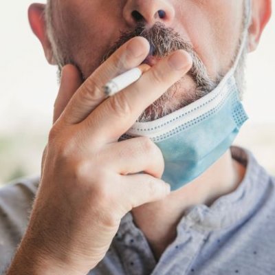 A close up of a man wearing a disposable mask over his chin, holding a cigarette to his lips. 